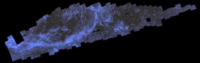 A wide, elongated black background with a long blueish patched-up field of stars, galaxies, and wispy clouds in the upper left diagonal of the image. This patchy mosaic consists of multiple almost square photographs, that are arranged in a shape that loosely resembles a thin, pixelated whale diving towards the bottom left of the image. The myriad of sparkling galaxies and stars in this mosaic crowd together to create the impression of a dark-grey field with frequent speckles of bright pastel yellow, red and blue dots of light at an even distribution. A streak of silver-blue diffuse clouds in a half-elliptical shape in the left half of the image, as well as a small round patch just right of the centre stand out particularly. At three spots right of the image centre there are small rectangular holes in the mosaic. Here, the pitch-black background is peaking through and stands in stark contrast to the sparkling image field.
