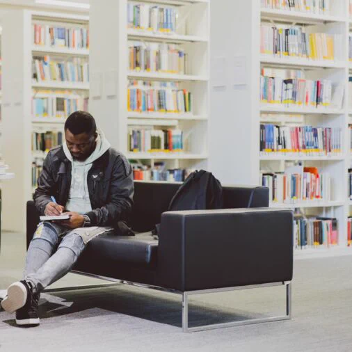 A student is sitting on a sofa in front of some library shelves full of books. The sudent is writing in a notepad on his knees.