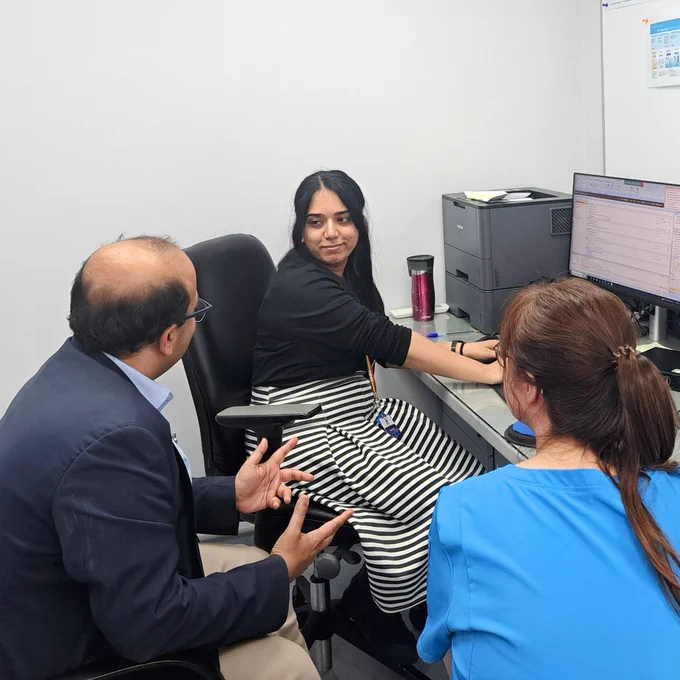 A photograph of Poonam sat at her desk inside of a consultation room. She is sat facing her colleagues.  
