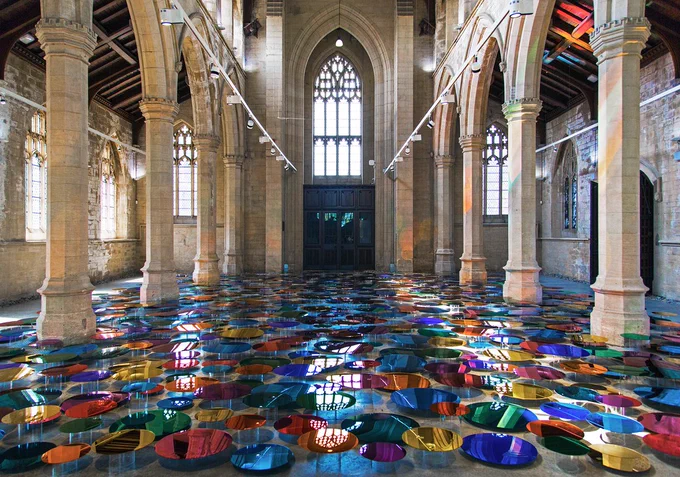 An installation of rainbow coloured mirrored discs displayed on the floor of a chapel.