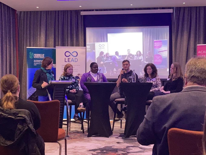 Five roundtable members sitting in front of several banners and a screen, which shows the live video stream. The chair is standing at a podium on the left.