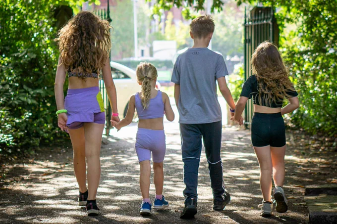 Four children walking out of Hesketh Park holding hands after a junior parkrun. Dappled sunlight coming through the trees