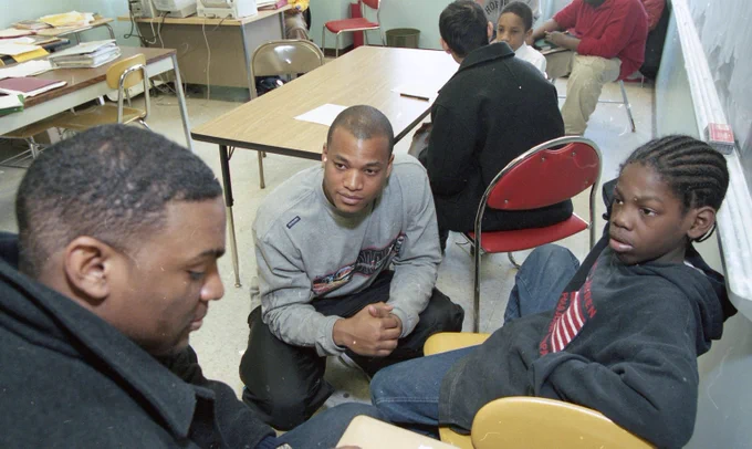 A crouched young Wes Moore crouches in front of a teen sitting in a chair as they listen to another young man in the foreground. 