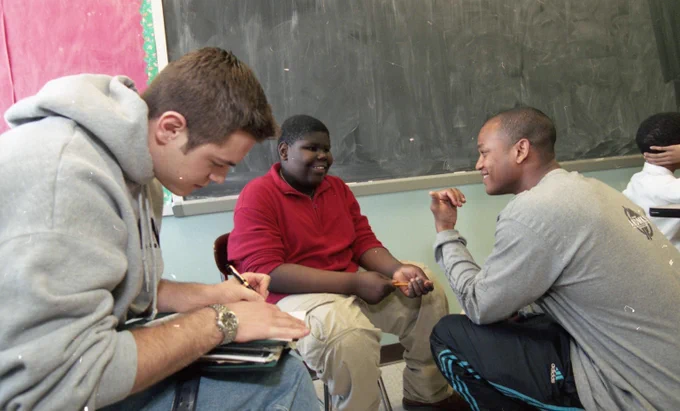 Wes Moore crouches as he and a teen share a laugh. Another young man sits nearby listening and writing. 