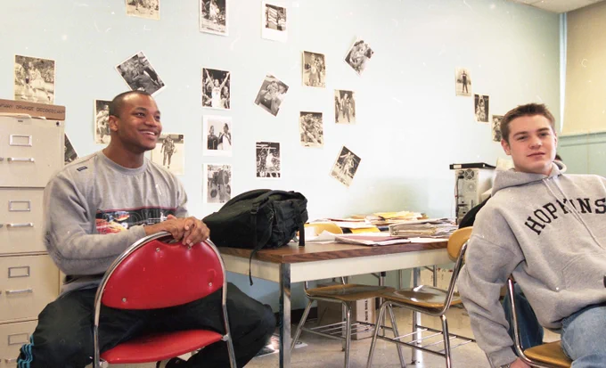 Wes Moore sits backwards in a chair smiling and another young man sits nearby wearing a Hopkins sweatshirt. 