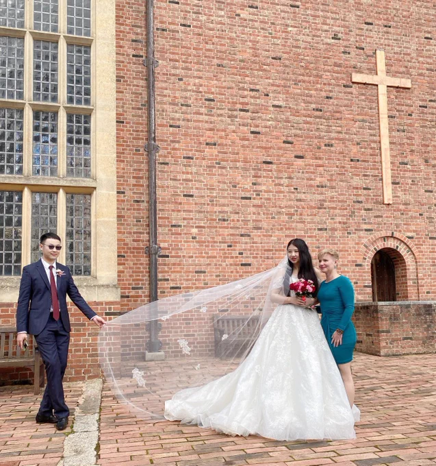 A bride, groom and guest stand outside a brick built chapel. There is a cross on the building and a large window made up of very small panes of glass.