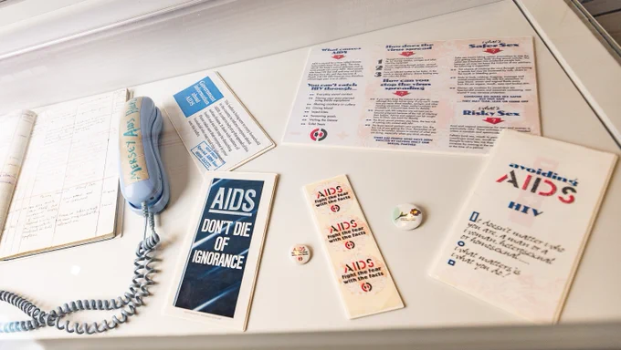 A glass display case containing a blue Merseyside AIDS Support Group telephone and open logbook, AIDS Don't Die of Ignorance leaflet and AIDS Fight the Fear with Facts badges, stickers and leaflets.