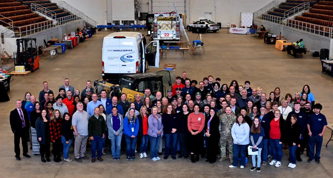 A group of about 50 people from local employers stands in the Veterans Memorial Coliseum surrounded by tables and equipment used in demonstrations. 