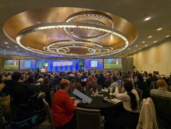 A large conference room with a stage in the middle accompanied by screens showing sign language interpreters and live captions. Nearly all the seats are full. 
