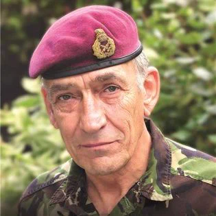 A British Army officer is facing the camera, wearing a maroon beret and a green camouflage uniform. In the background is green foliage.