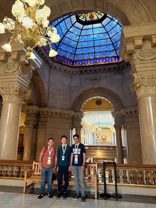 Three individuals stand smiling inside the ornate Indiana Statehouse. Above them is a grand stained-glass dome with blue and gold accents. Large columns and decorative arches frame the background, with a golden chandelier hanging on the left. The interior features a blend of detailed stonework and golden railings.

