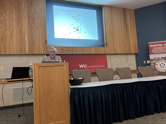 Keith Hobson speaks from behind a podium with a projected slide that reads “Some reflections on SIA and IsoEcology.”