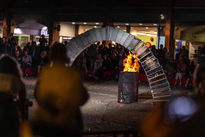 Outdoors at night in a public city square. A crowd of people stand and sit around an arch made of concrete and ice blocks. A fire burns underneath it, melting the ice.