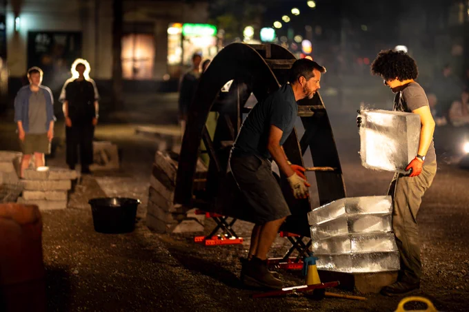 Two performers are building arch while four more sing, facing them. Nao lifts a heavy, slippery ice block, while Seth bends his knees, bracing and reaching out to help her position it. Verity, Ellian, Phil and Aminita stand on the opposite side of the arch, watching them and singing.