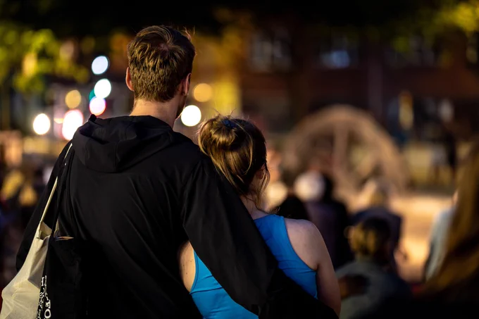 Two audience members watch the performance, embracing and leaning against each other in the growing darkness.