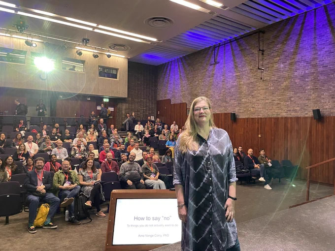 Aino standing on stage presenting a slide titled "How to say 'no'", with an audience looking on in a room with purple lighting.