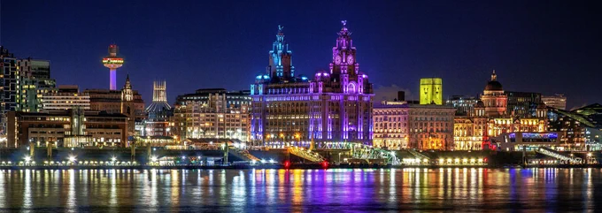 The image shows the Liverpool skyline at night. The iconic Liver Building is lit up purple. The lights are reflected in the Mersey.