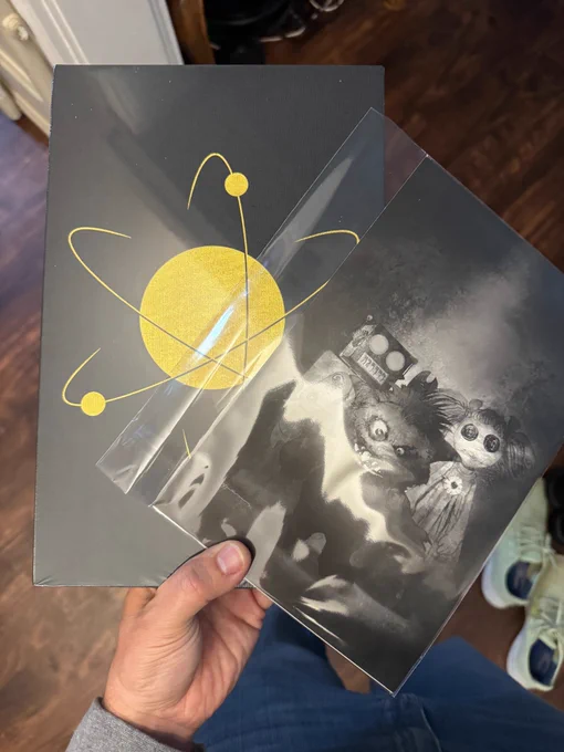 A white hand holds a Lab Press wrapped book and lithograph of ESSENTIALS cover art featuring Snuggles. 