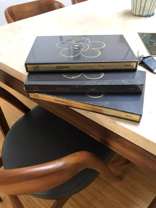 A stack of newly delivered copies of ESSENTIALS are stacked at an angle on a white quartz table top. There are car keys above and to the right of the books, and a portion of 1 chair with wooden arms and a black cushion in the background.