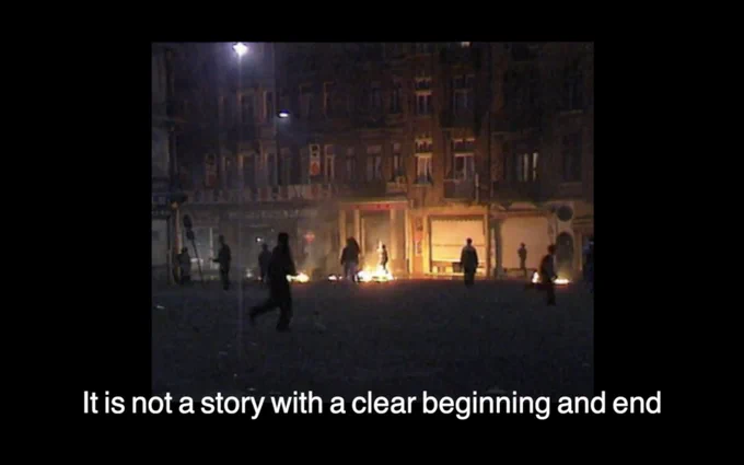 A city street at night. The shops are shuttered, and there is a fire on the ground in front of one of them. In the foreground, a figure dressed in dark clothes appears to be running. Others stand in the background. Beneath the image is a subtitle that reads "It is not a story with a clear beginning and end."