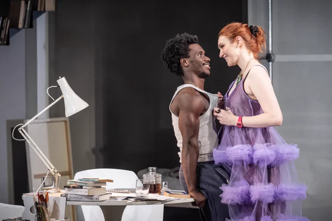 Michael Ahomka-Lindsay and Eleanor Tomlinson in Reverberation. They're close together against a desk. She wears a purple frill dress and he a grey vest. They're looking at each other adoringly.
