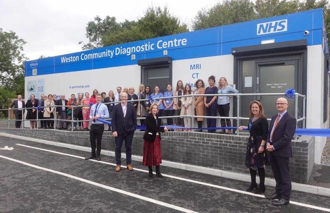 A large group of people are gathered in front of the newly opened Weston Community Diagnostic Centre. In the foreground, UHBW CEO Maria Kane is cutting a blue ribbon.