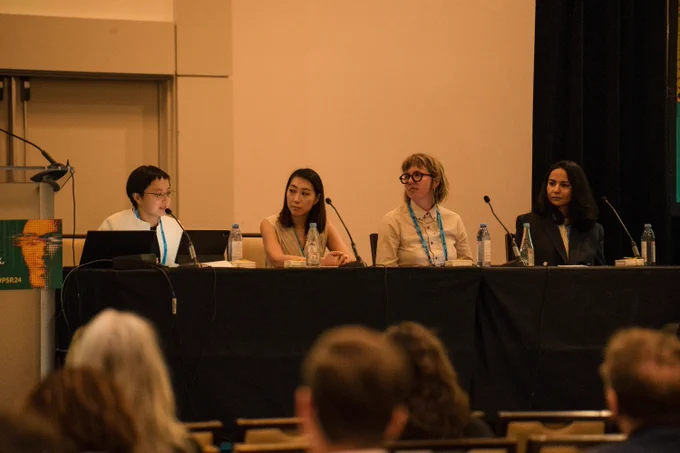 Four panelists are seated at a table during a conference discussion. They appear engaged and attentive, with microphones in front of each speaker. The audience in the foreground listens intently.