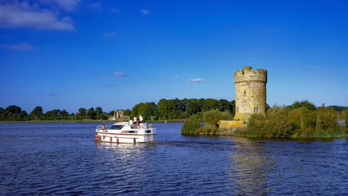 A photo of a white boat on a body of water, close to a small piece of land containing green foliage and a small, stone round tower. In the background is the shore, covered in green trees, and a blue sky with clouds.