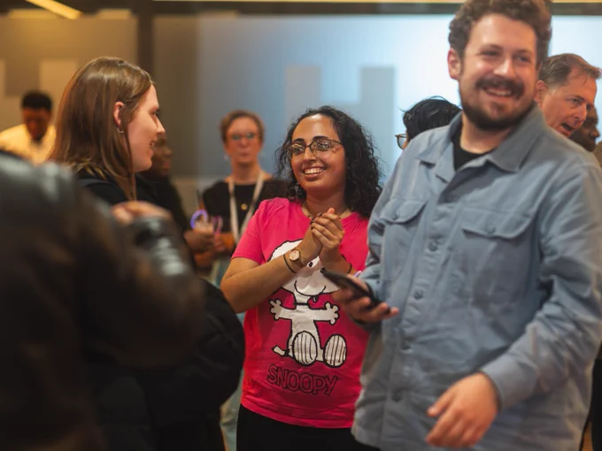 A group of people socializing at an event, one person wearing a pink Snoopy T-shirt and another in a blue denim shirt, both smiling and engaged in a conversation.