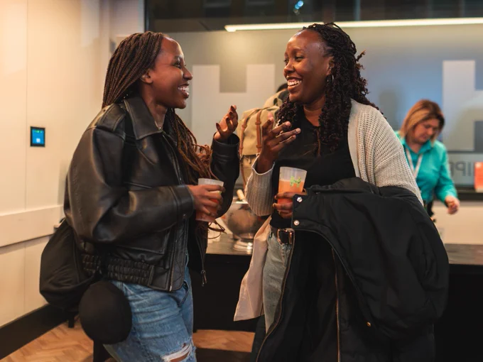 Two people laughing and holding drinks at a social event, inside a room with modern decor.