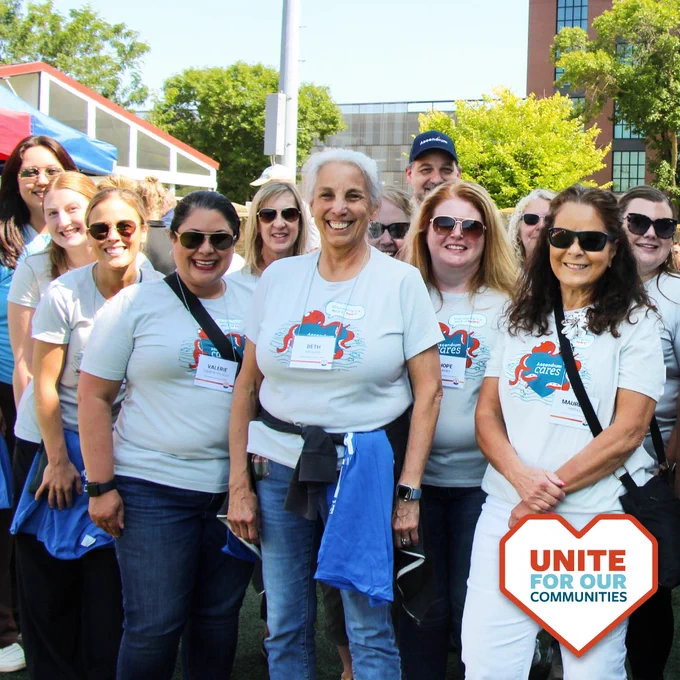 A group of twelve Ascendium employees smile during an outdoor volunteer opportunity. A logo in the lower-right corner reads, "Unite for Our Communities" and is outlined by a heart shape.