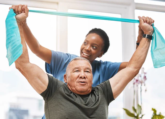 A physical therapist assists an older adult with an exercise using a resistance band in a bright, well-lit room.