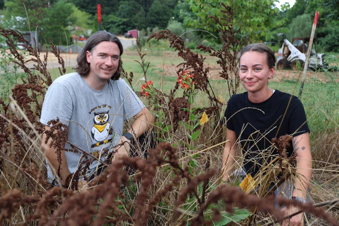Two individuals working in a field with dried plants, one wearing a T-shirt with an owl graphic, both smiling at the camera.