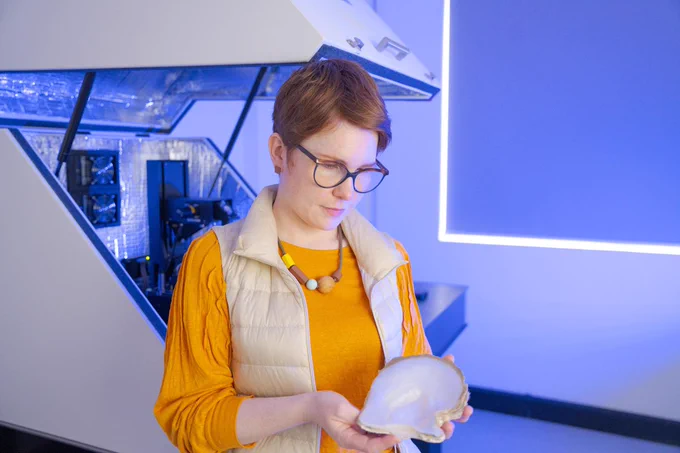 Dr. Otter studies a piece of a marine shell while standing in the Photo-induced Force Microscopy lab, with a large metallic piece of equipment behind her. Photo: Nic Vevers/ANU