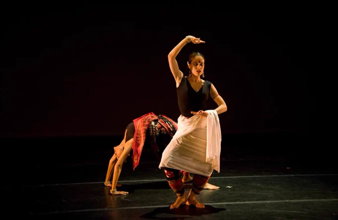 A woman puts her right hand over her head as she dances, while another dancer makes a U-shape on the stage with her midsection pointed upward and hands and feet down.