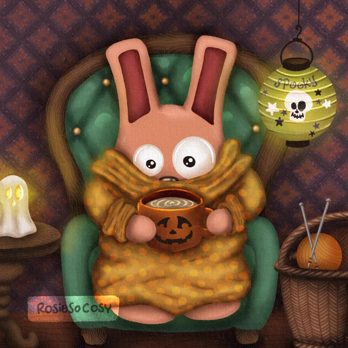 It’s an illustration of a pink Freezer Bunny with dark pink ears. Freezer Bunny is wearing an ochre yellow dotted snuggly blanket type sweater, holding an orange pumpkin mug with what looks like a latte. Freezer Bunny is seated on a teal coloured armchair with a diamond tuffed pattern on the back, and a wooden frame and legs. On the left is a round end table with a ghost candle holder. On the right is a wicker basket on the floor with little balls of yarn and knitting needles. Above it hangs a green paper lantern with a skull, stars and in purple the word “spooky”