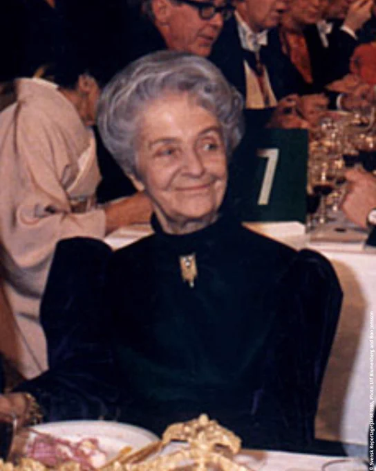 Rita Levi-Montalcini sits at the Nobel Prize Banquet, dressed in a black outfit and wearing a golden necklace, with other attendees in the background.