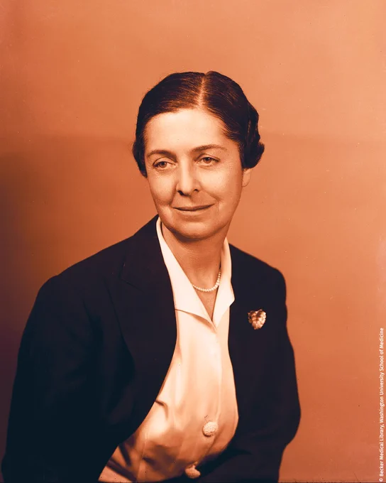 Portrait of Rita Levi-Montalcini, dressed in a dark blazer and light blouse, wearing a brooch, looking at the camera with a slight smile.