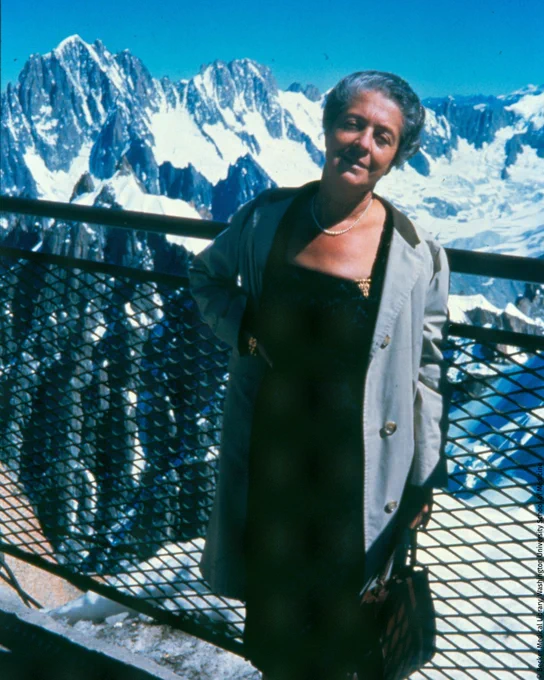 Rita Levi-Montalcini stands in front of snowy mountain peaks, wearing a dark dress and a light coat, with a serene expression.