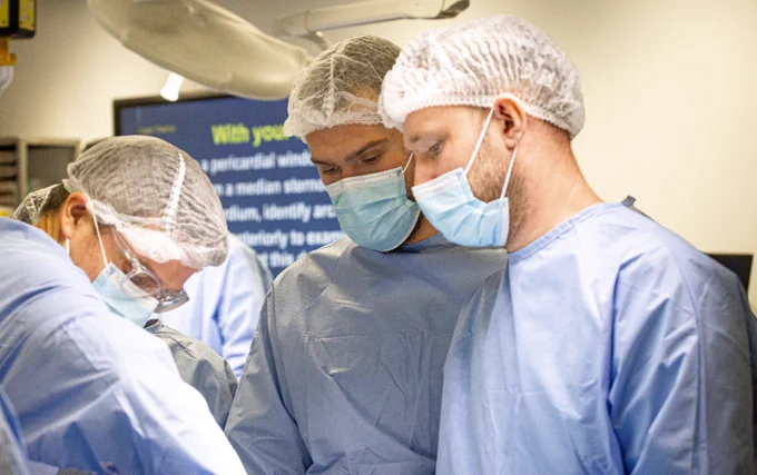 Surgeons intently observe another surgeon at work. In the background a training slide of surgical procedure information is on a large screen in a surgical training simulation. 