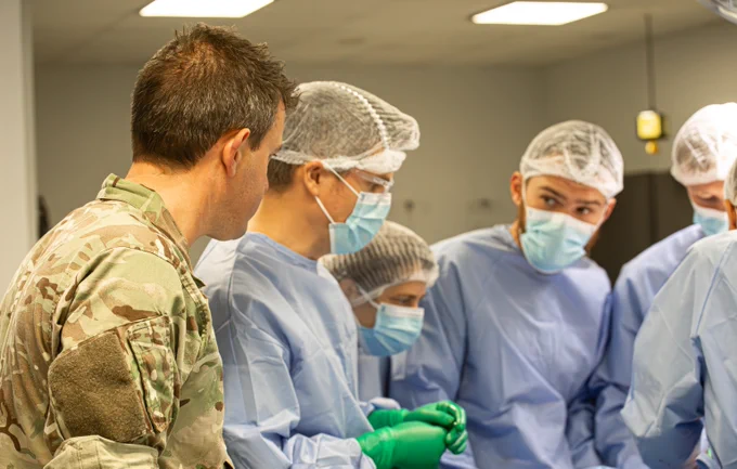A British military surgeon in uniform provides instruction to a mixed surgical team of Ukrainian and UK doctors wearing scrubs in a surgery simulation.