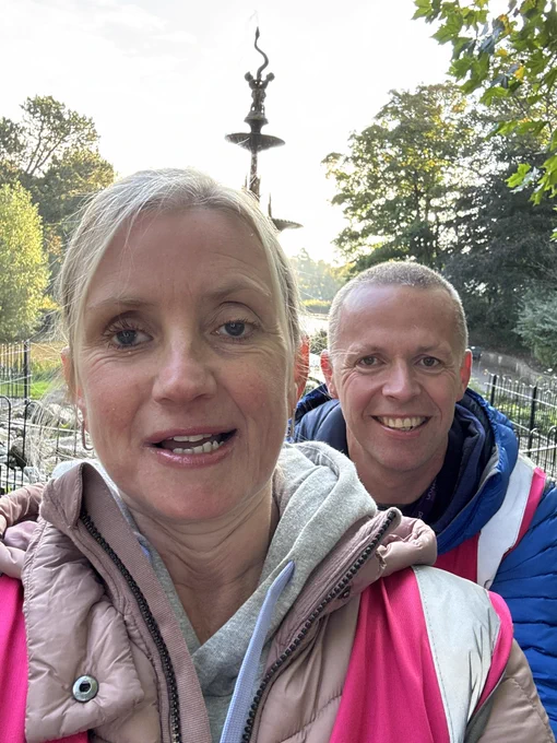 The image shows a close-up selfie of a woman and a man outdoors. The woman is in the foreground, smiling slightly, with light blonde hair pulled back. She is wearing a light pink puffer jacket over a gray hoodie. The man is standing slightly behind her, smiling broadly. He has short-cropped hair and is wearing a blue jacket with a pink vest over it. In the background, there is a decorative fountain with multiple tiers and a sculptural element on top. The setting appears to be a park or garden with trees and greenery visible in the background. The lighting suggests it might be early morning or late afternoon.