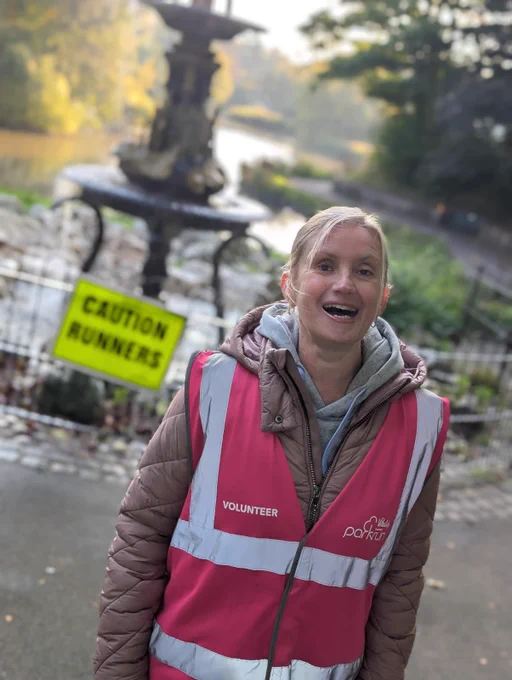 The image shows a woman standing outdoors in a park. She is wearing a bright pink high-visibility vest with reflective stripes. The vest has the word "VOLUNTEER" printed on it, along with a logo that says "parkrun." Underneath the vest, she is wearing a pink quilted jacket and a grey hoodie. She is smiling and appears to be happy. In the background, there is a fountain and a body of water, with trees and greenery surrounding the area. There is also a yellow sign with black text that reads "CAUTION RUNNERS" positioned near the fountain. The setting suggests a park or natural area, possibly during an event like a run or walk.