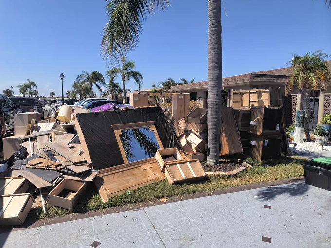 A scene of extensive debris and damage outdoors, possibly after a storm, with broken wooden structures and scattered household items around a palm tree.