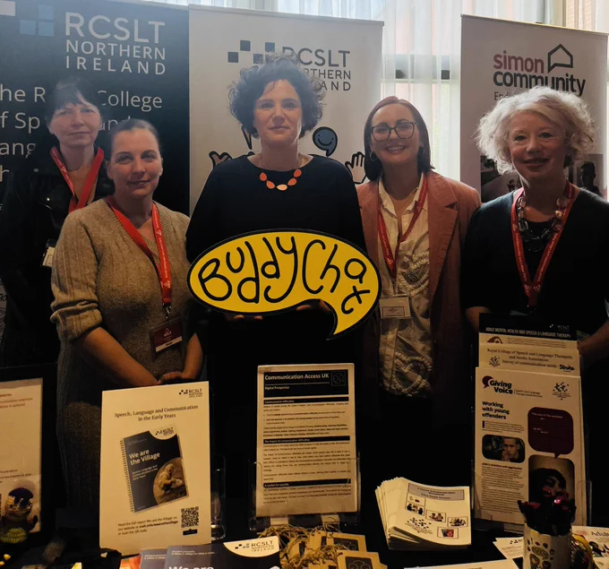 SDLP MP Claire Hanna engages with RCSLT NI representatives. Standing behind table holding Buddy Chat prop.