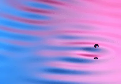 A small liquid droplet hovers above a rippled, light pink and blue fluid surface that takes up the entire image. The droplet is reflected on the surface, and it appears to be at a standstill as the surface ripples interact with it.