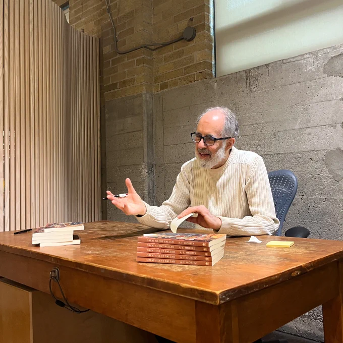 A photo of Al Moritz taking a mid-book signing break to chat with someone just out of view. A stack of copies of Great Silent Ballad is on the table in front of him.