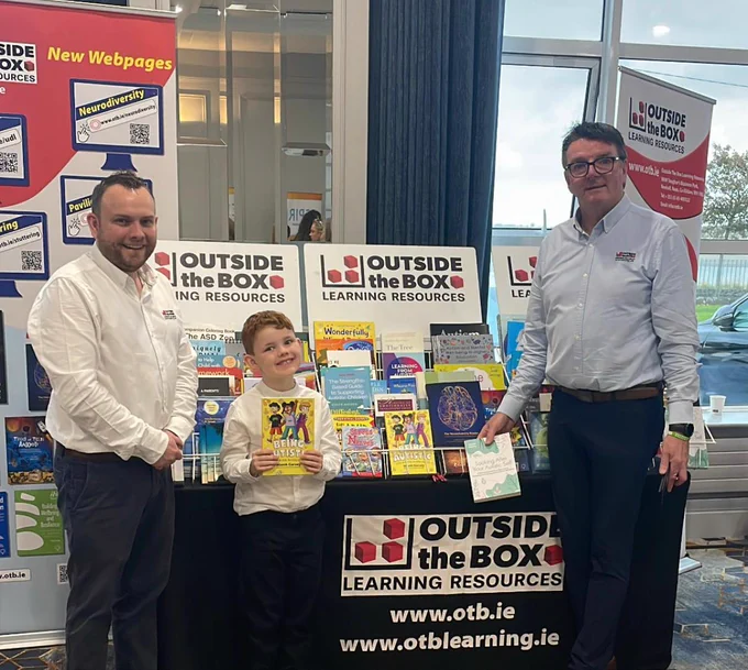 Photo of book stall by Outside the Box Learning Resources, with 2 men, and one boy who is holding my book "Being Autistic (and what that actually means)"
