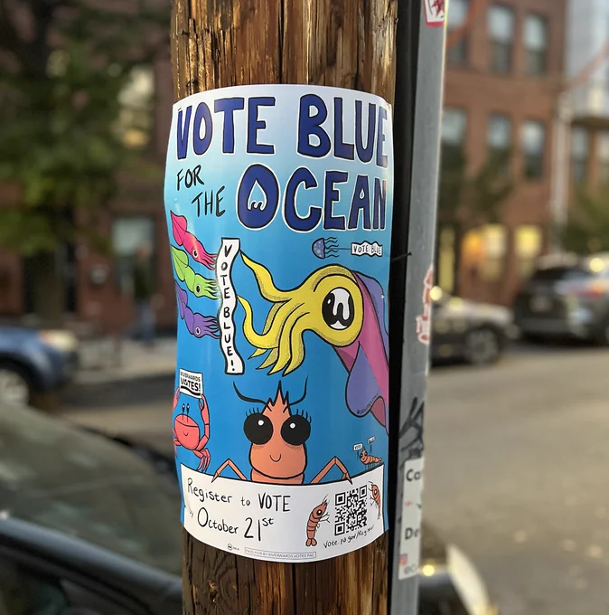 a poster on a pole with a bunch of sea creatures that says Vote Blue for the Ocean, with squid holding up a sign that says vote blue, and a jellyfish dragging a banner that says Vote Blue and a little shrimp holding up two signs, each of which say Vote andBlue, The big shrimp with eyelashes is saying Register to Vote by October 21 and a QR code to register