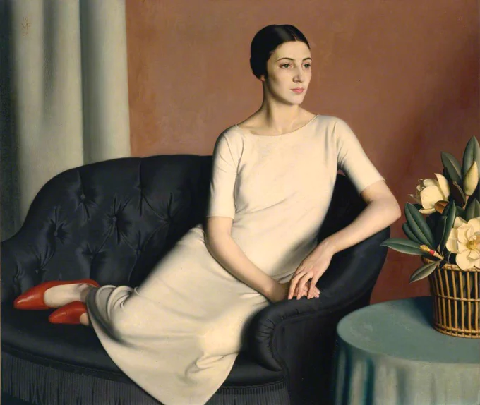 A woman sits on a dark sofa with her legs up, she wears a nude coloured dress and bright red shoes. She's looks to the right. There's a table with white flowers on.